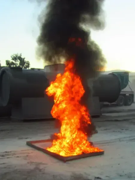 A bright orange fire burns in a rectangular metal tray, producing thick black smoke, set outdoors near industrial tanks.