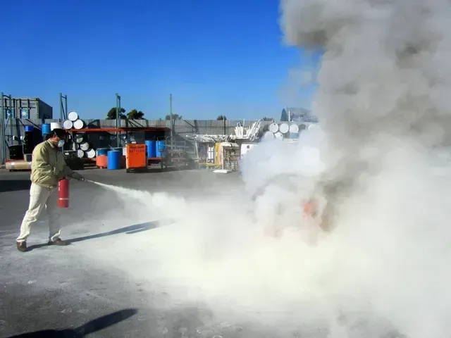 A person in tan work clothes uses a fire extinguisher to spray a white powder onto a small fire in an industrial yard.