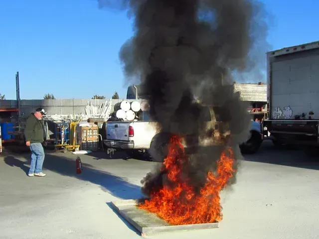 A person stands near a fire burning in a metal tray, producing a large cloud of thick black smoke in an outdoor yard.