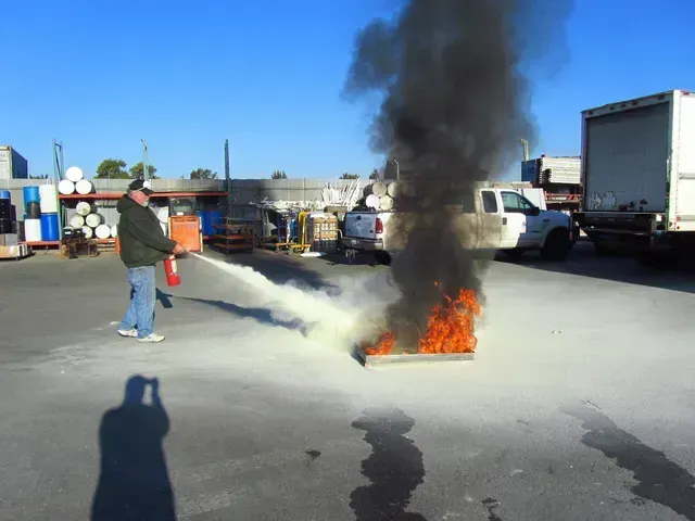 A person wearing a dark jacket and jeans uses a red fire extinguisher to put out a small, intense fire on a paved lot.