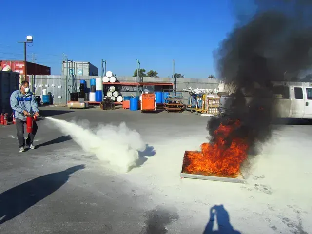 A person in a protective mask uses a fire extinguisher to put out a fire in a metal pan outdoors in an industrial yard.