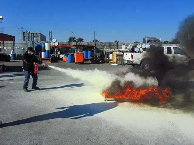 A person uses a fire extinguisher to put out a small fire on asphalt in an industrial area.