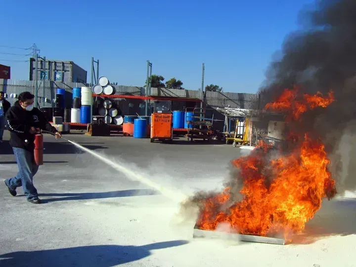 A person uses a fire extinguisher to put out a fire in a metal pan in an industrial outdoor setting.
