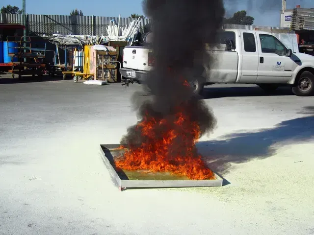 A small rectangular pan filled with burning fuel emits flames and thick black smoke in an industrial parking lot.