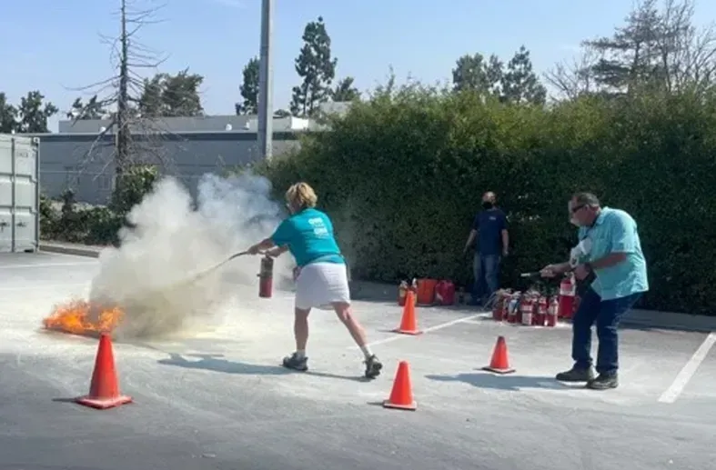 Two people practice using fire extinguishers on a small outdoor fire in a parking lot marked with orange safety cones.