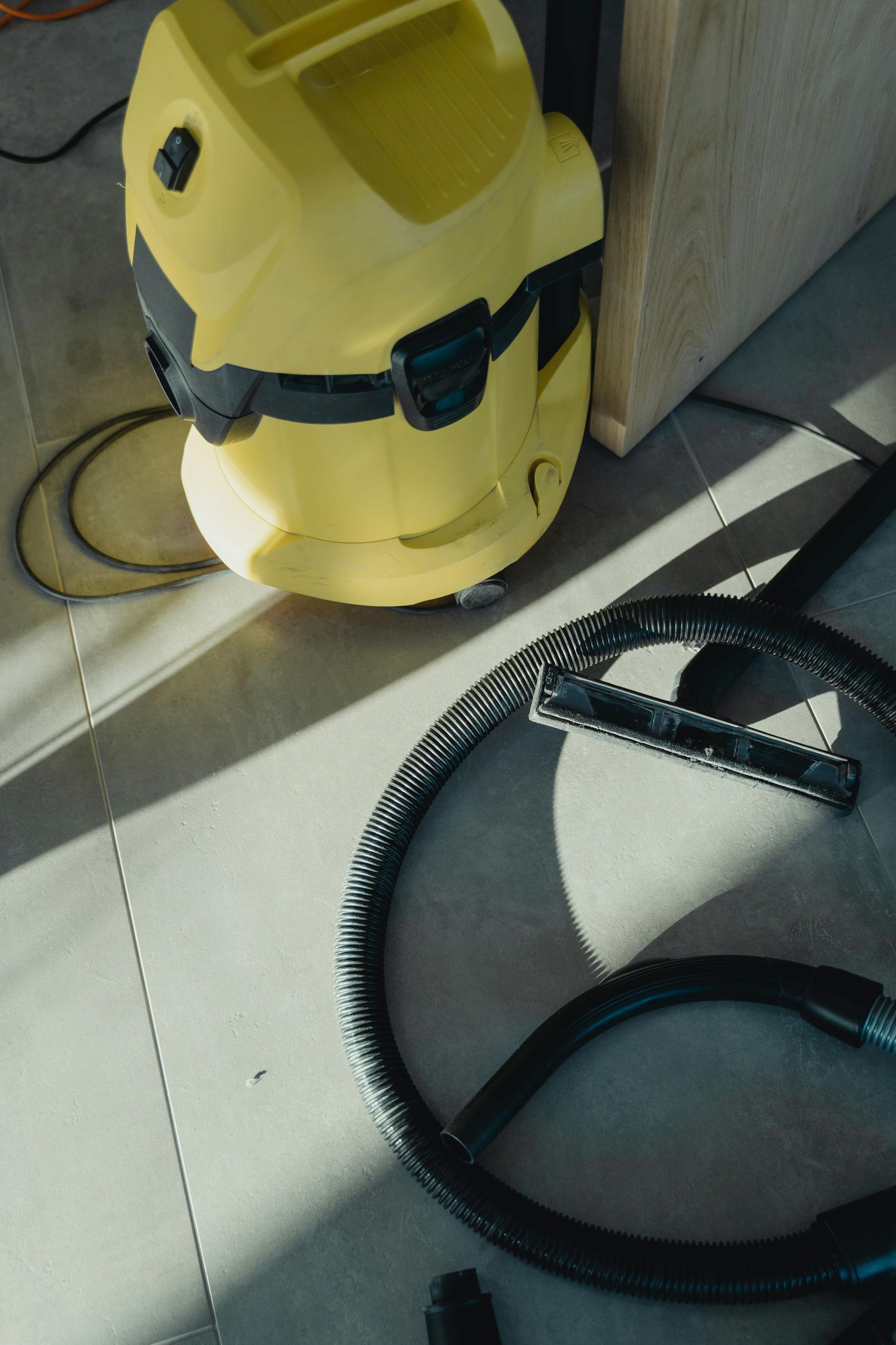 Yellow shop vacuum with black hose on a sunlit floor near a wall