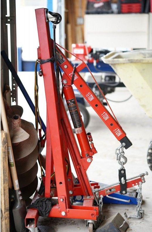 A red crane is sitting in a garage next to a wheelbarrow.