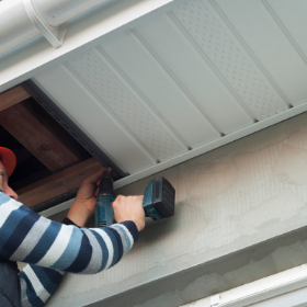 A person uses a power drill to install white perforated soffit panels under a roof eave.
