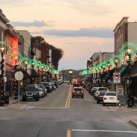 A quiet town street at dusk, lined with holiday lights and parked cars, under a soft pastel sky.