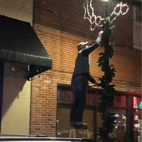 A person stands on a step ladder to arrange a holiday light display on a vertical pole outside a brick building.