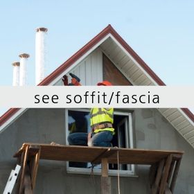 A construction worker in a high-visibility vest kneels on scaffolding to install soffit and fascia on a house gable.