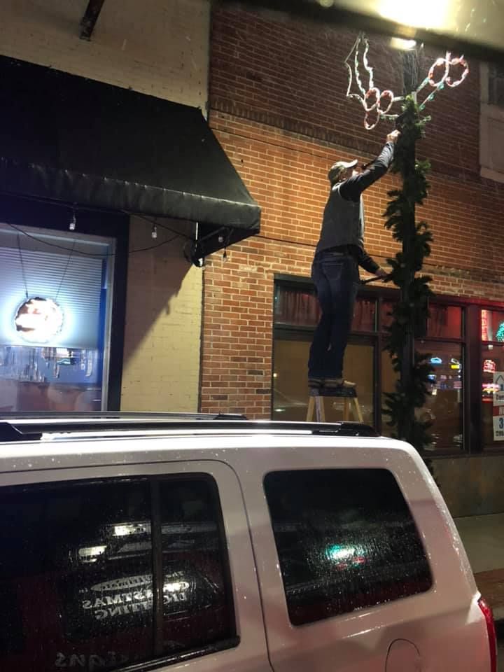 A person stands on a ladder on top of a car to decorate a post with greenery and light-up holly ornaments at night.