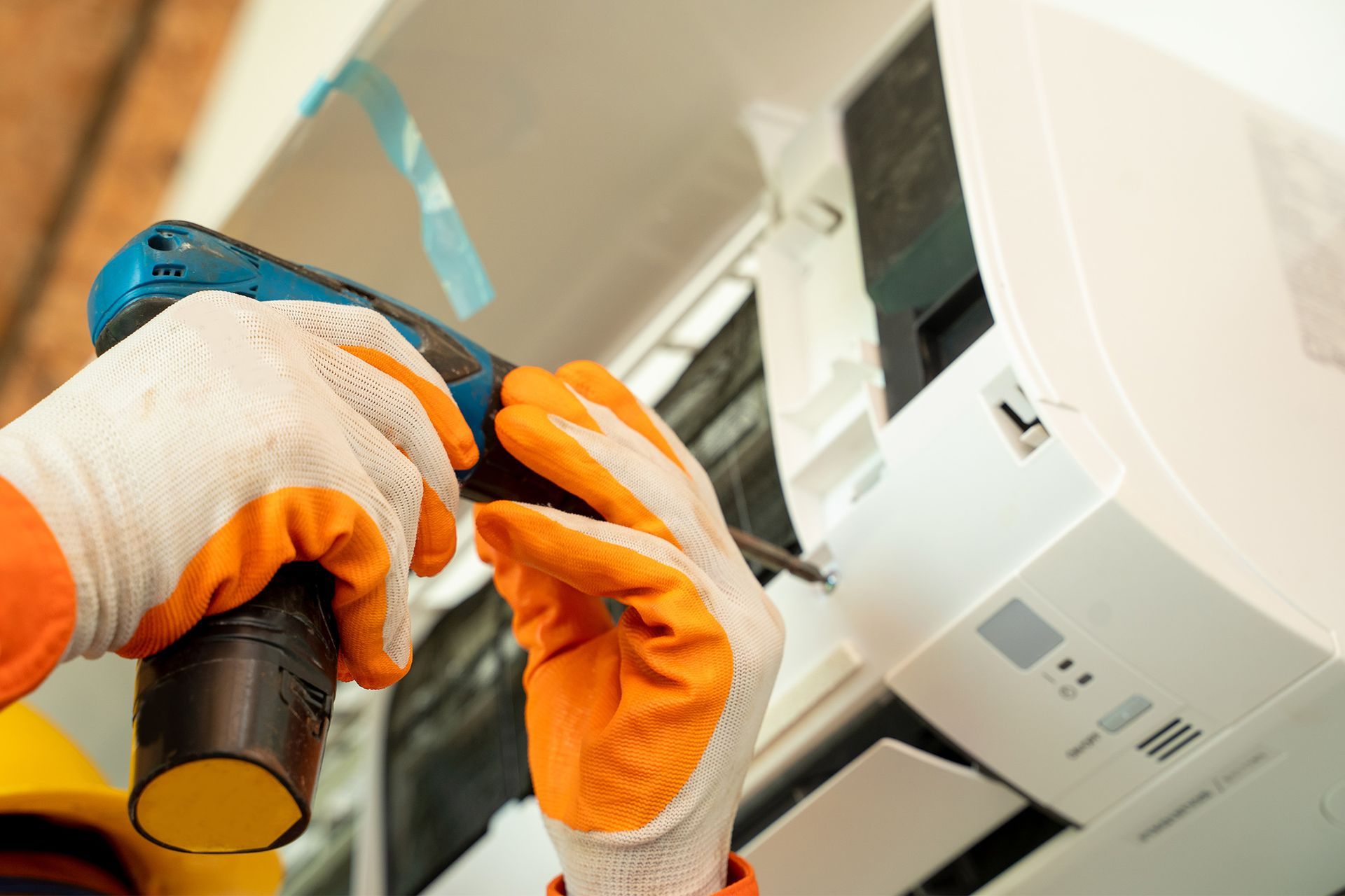 A man wearing orange gloves is working on an air conditioner with a drill.