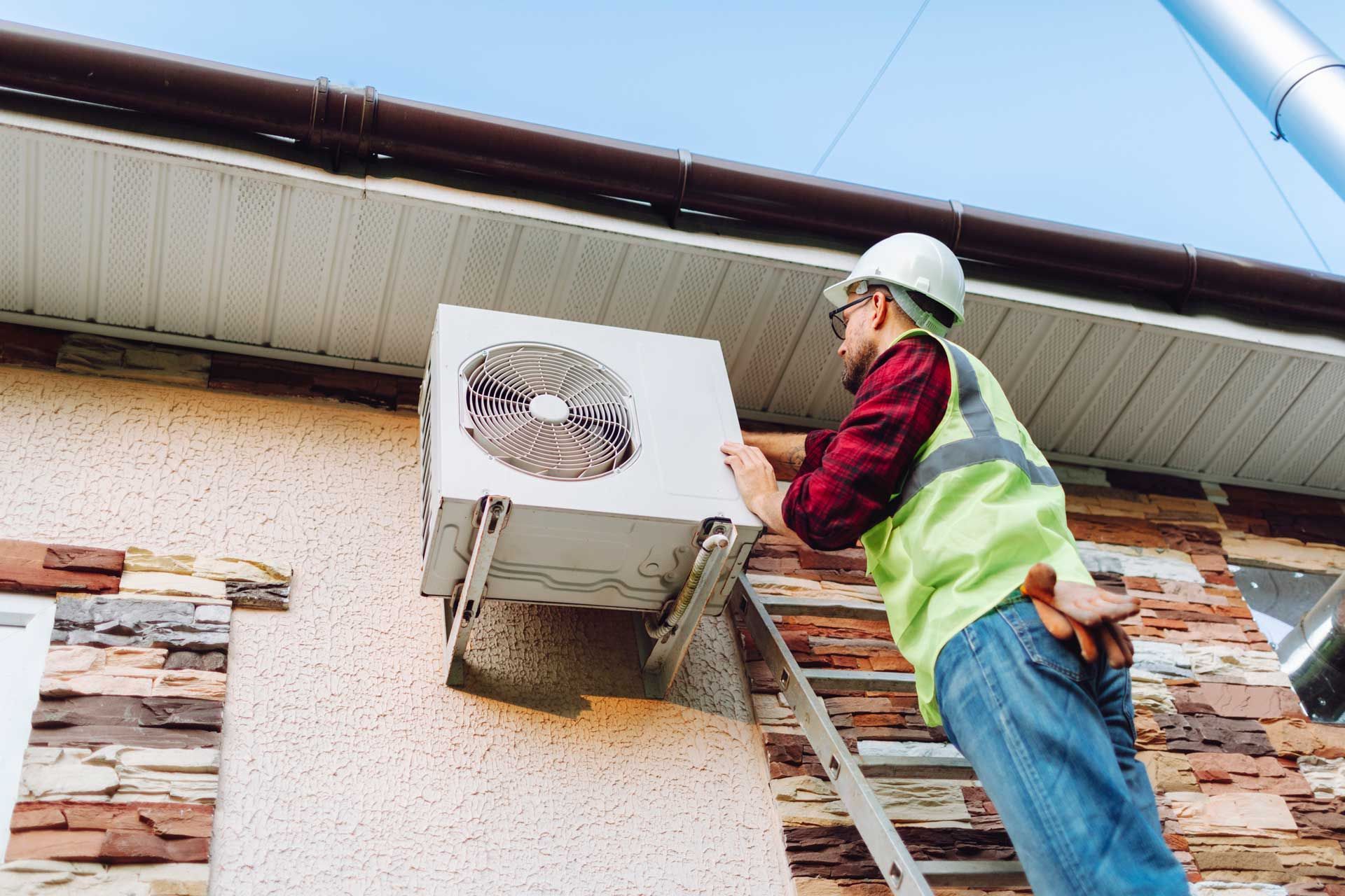 A man is standing on a ladder installing an air conditioner on the side of a building.