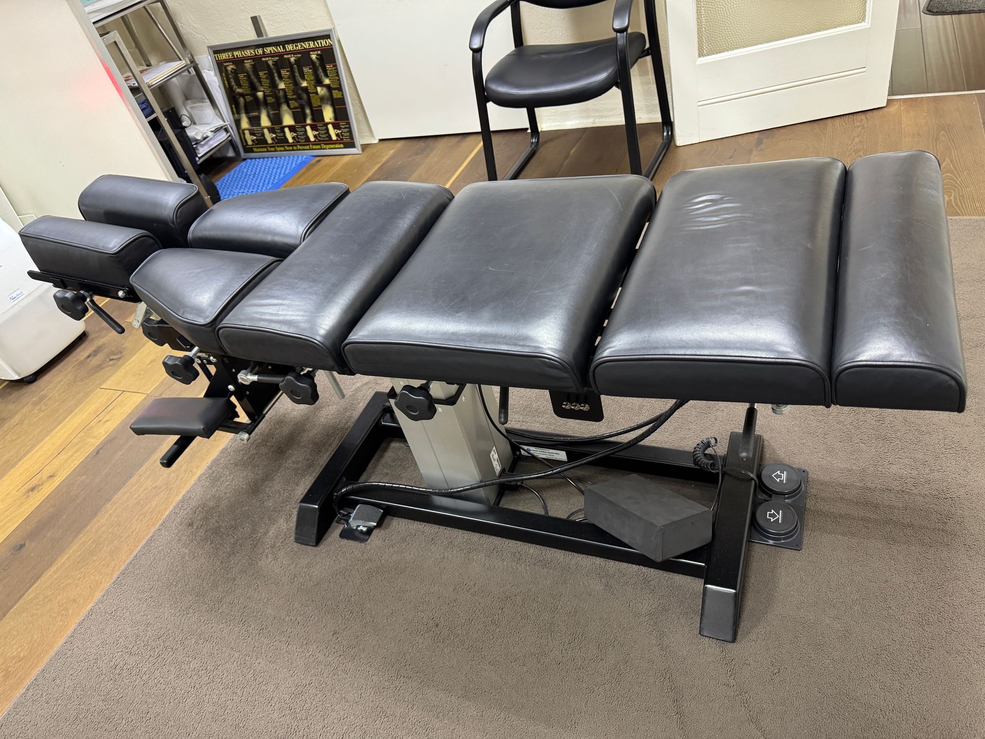 Black chiropractic adjusting table in a room with a chair and trophies.