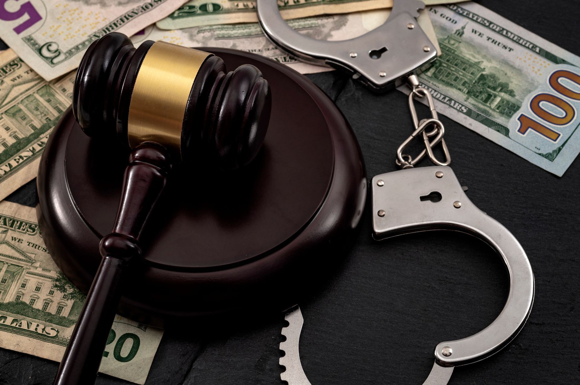 A wooden judge's gavel resting on a sound block next to metal handcuffs and scattered bills.