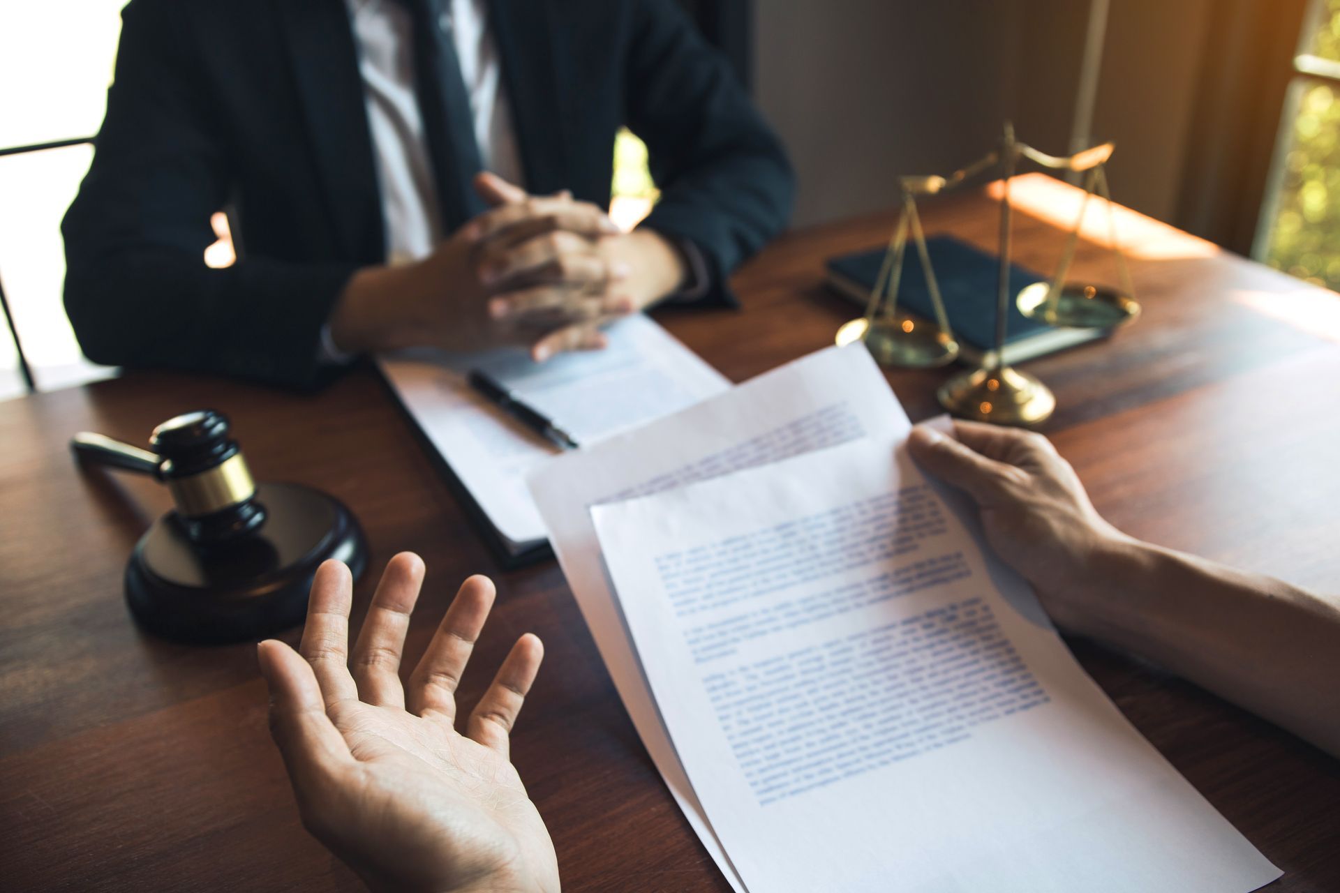 A lawyer sits at a desk with a gavel and scales of justice as a client discusses legal documents.