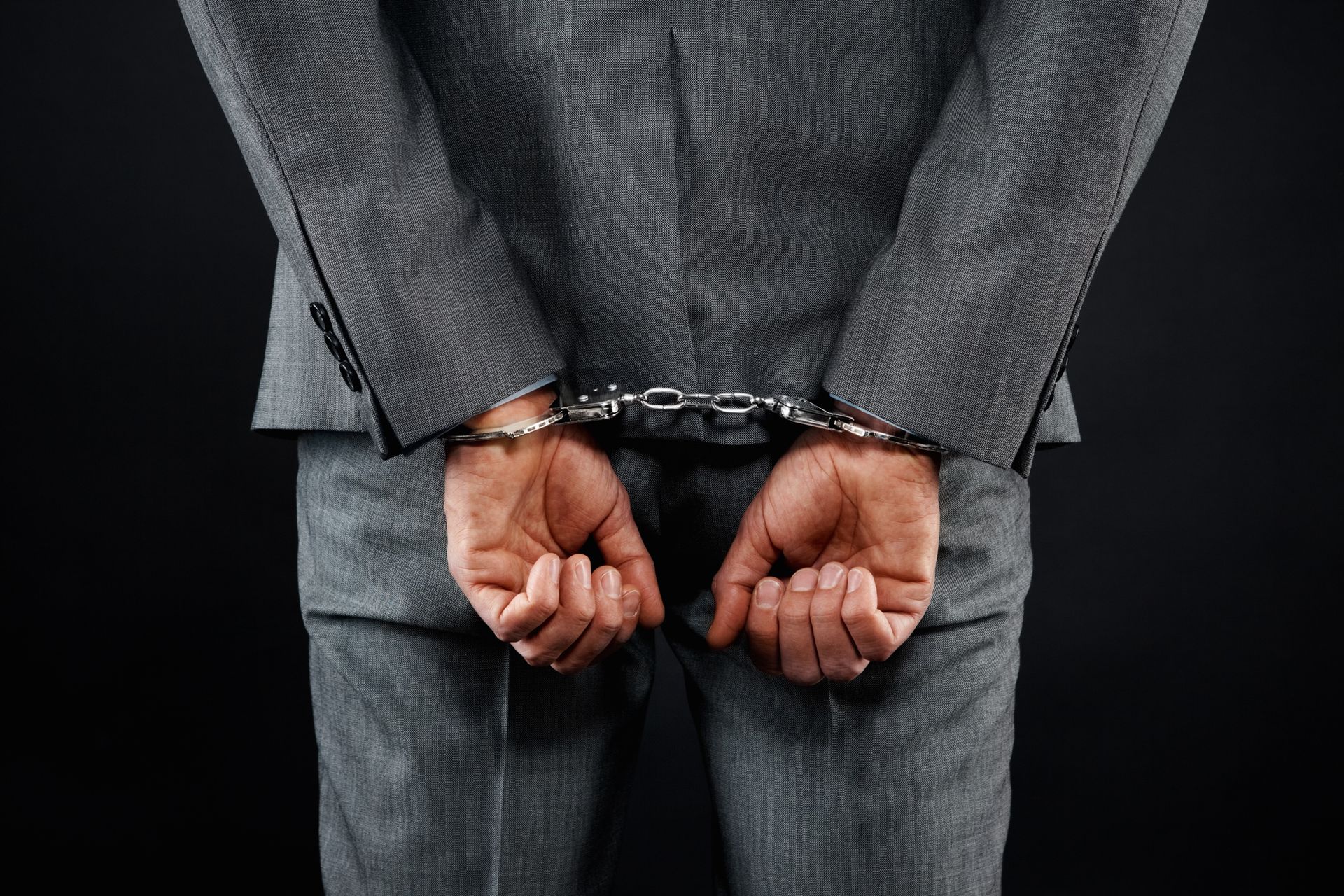 A person in a suit, seen from the back, has hands cuffed behind their back.