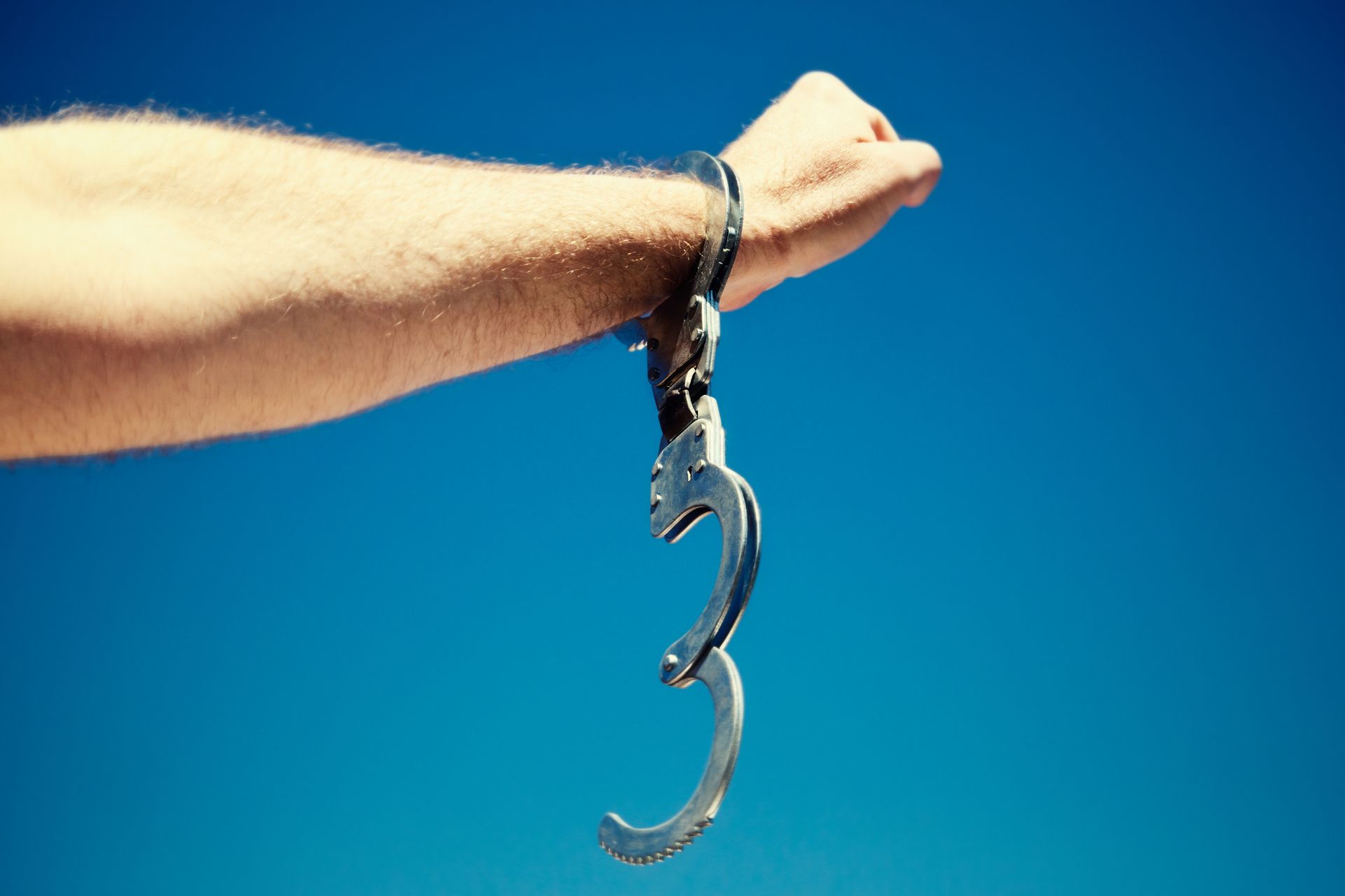 Hand with open handcuffs against a bright blue sky.