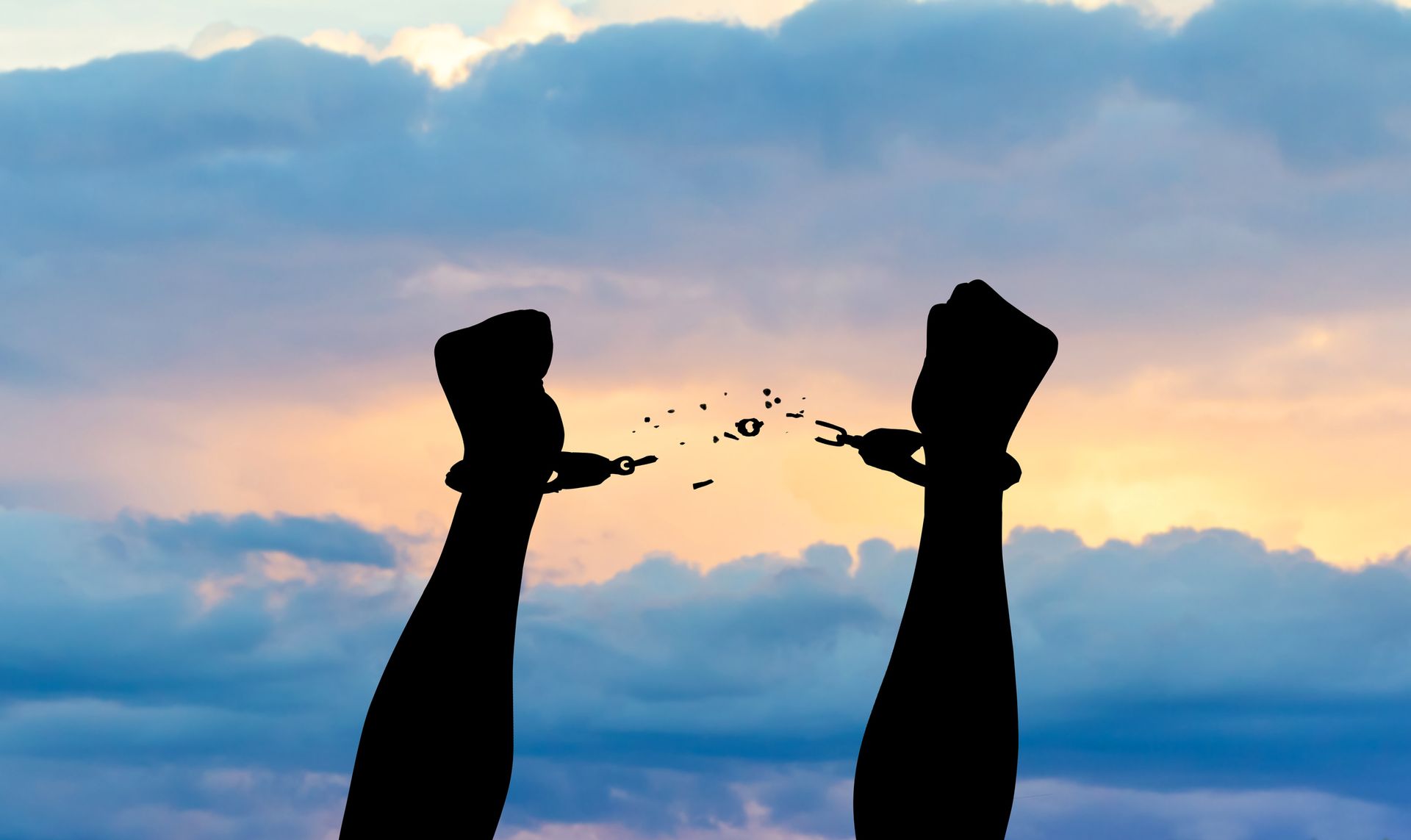 Silhouette of hands breaking shackles against a sunset sky, symbolizing freedom.