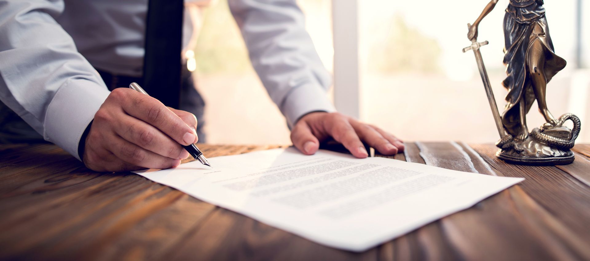 A person signing a document on a wooden table, with a Lady Justice statue in the background.