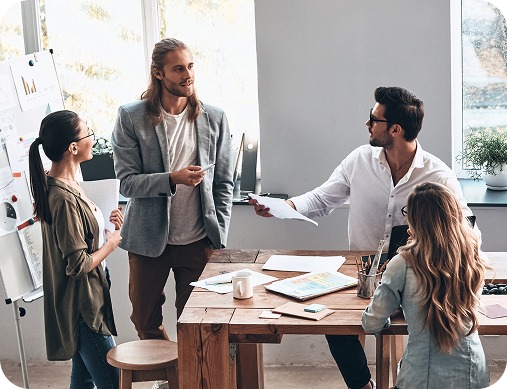People in a business meeting. Three stand near a whiteboard. Two others sit at a wooden table with papers.