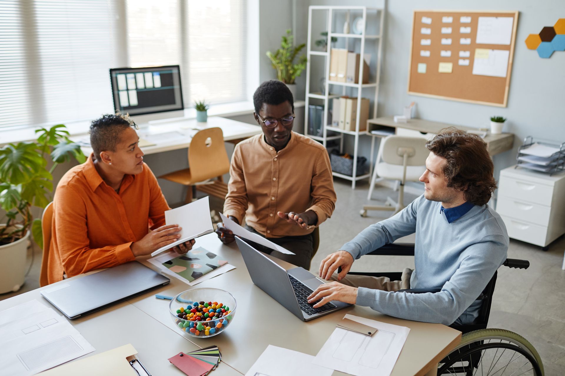 Three people collaborating at a table in an office, one using a laptop, one in a wheelchair.