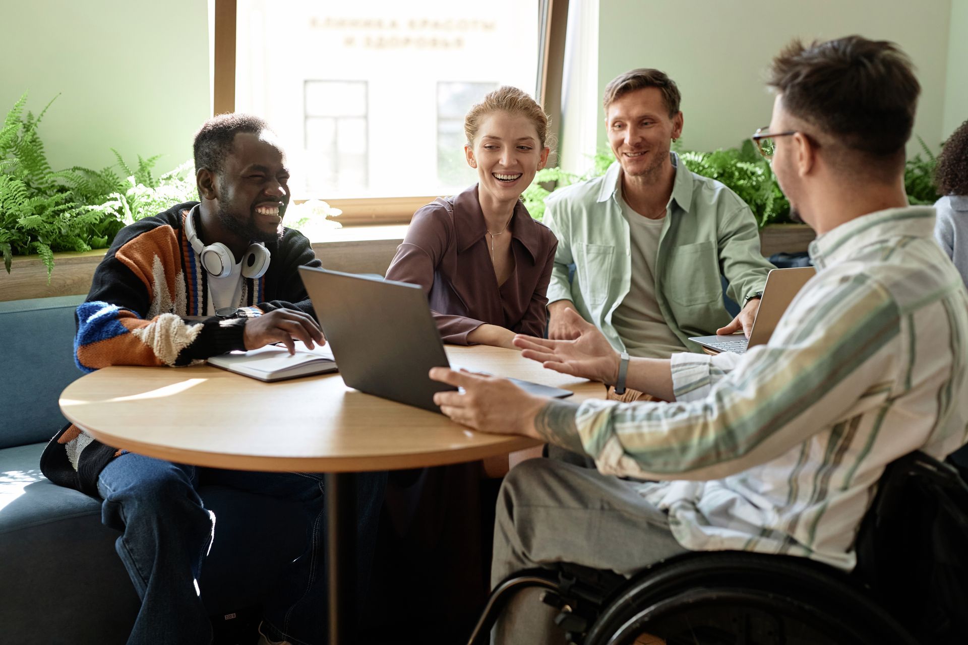 Four people laugh while gathered around a round table, one using a laptop, one in a wheelchair.