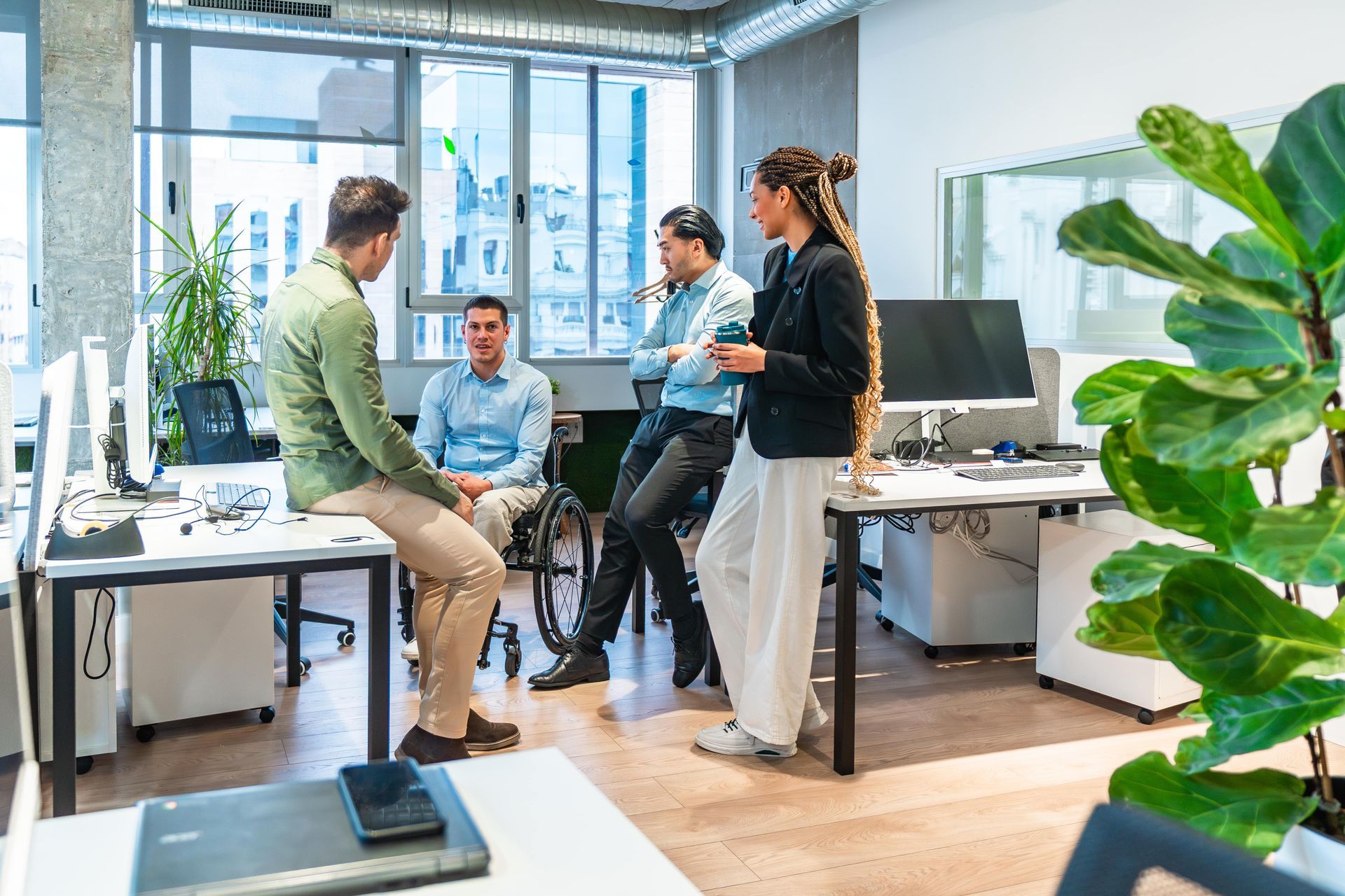 Four people in an office. One in a wheelchair, others standing. White desks, large windows, indoor plant.