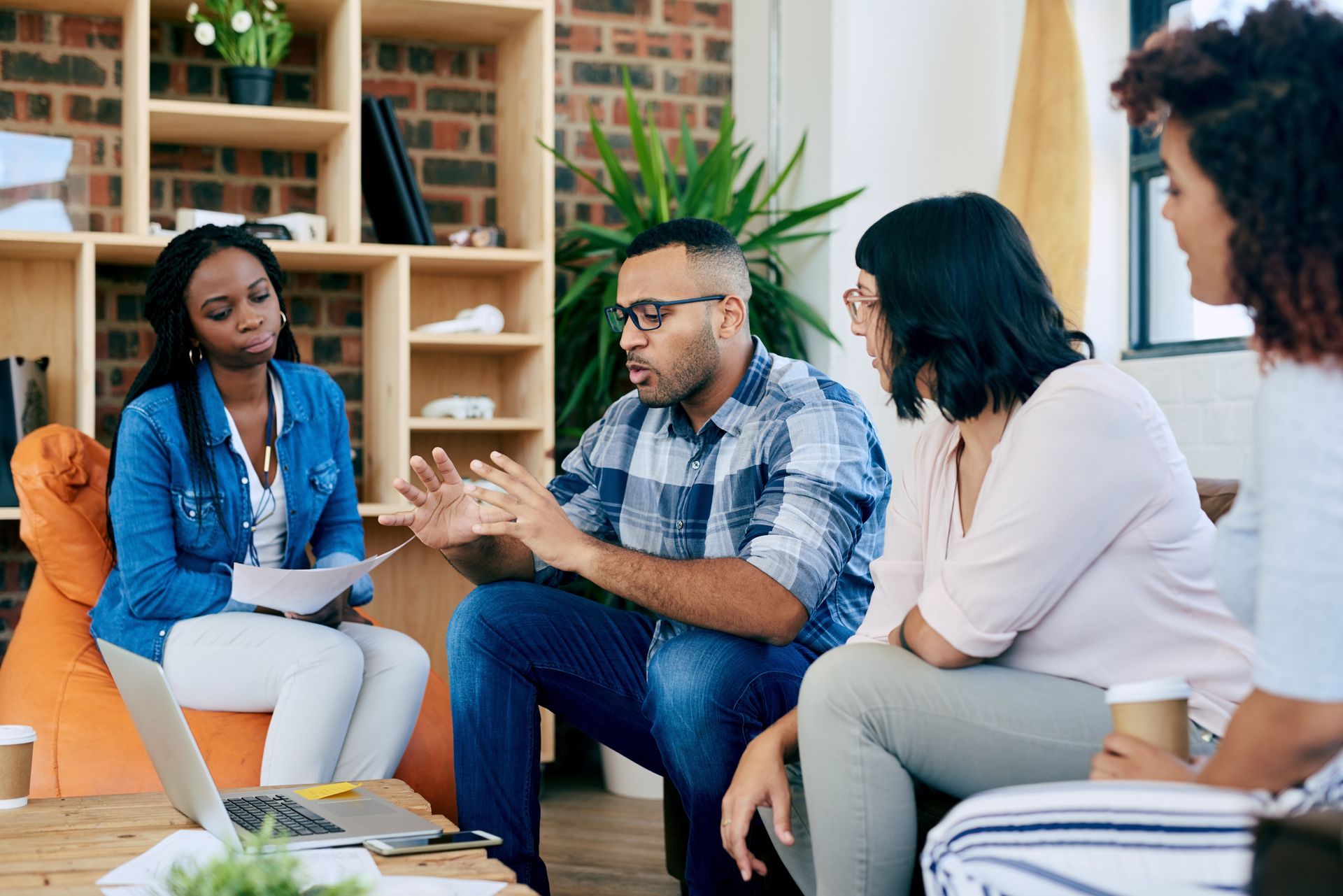 Group of people in discussion, sitting in a living room. Man gestures with hands, others listen with thoughtful expressions.