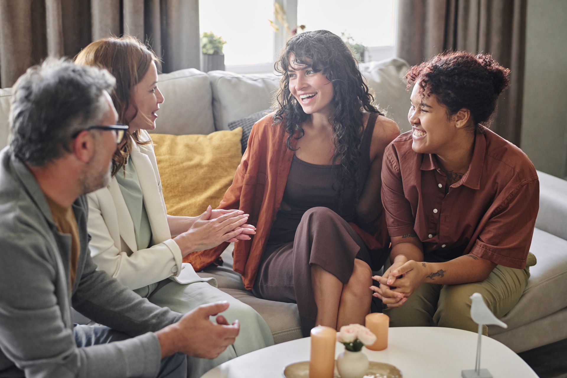Four people seated on a couch, talking and smiling. Interior room with coffee table and candles.