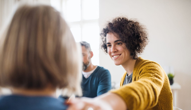 Person with curly hair smiles, placing hand on another's shoulder, in a counseling session.