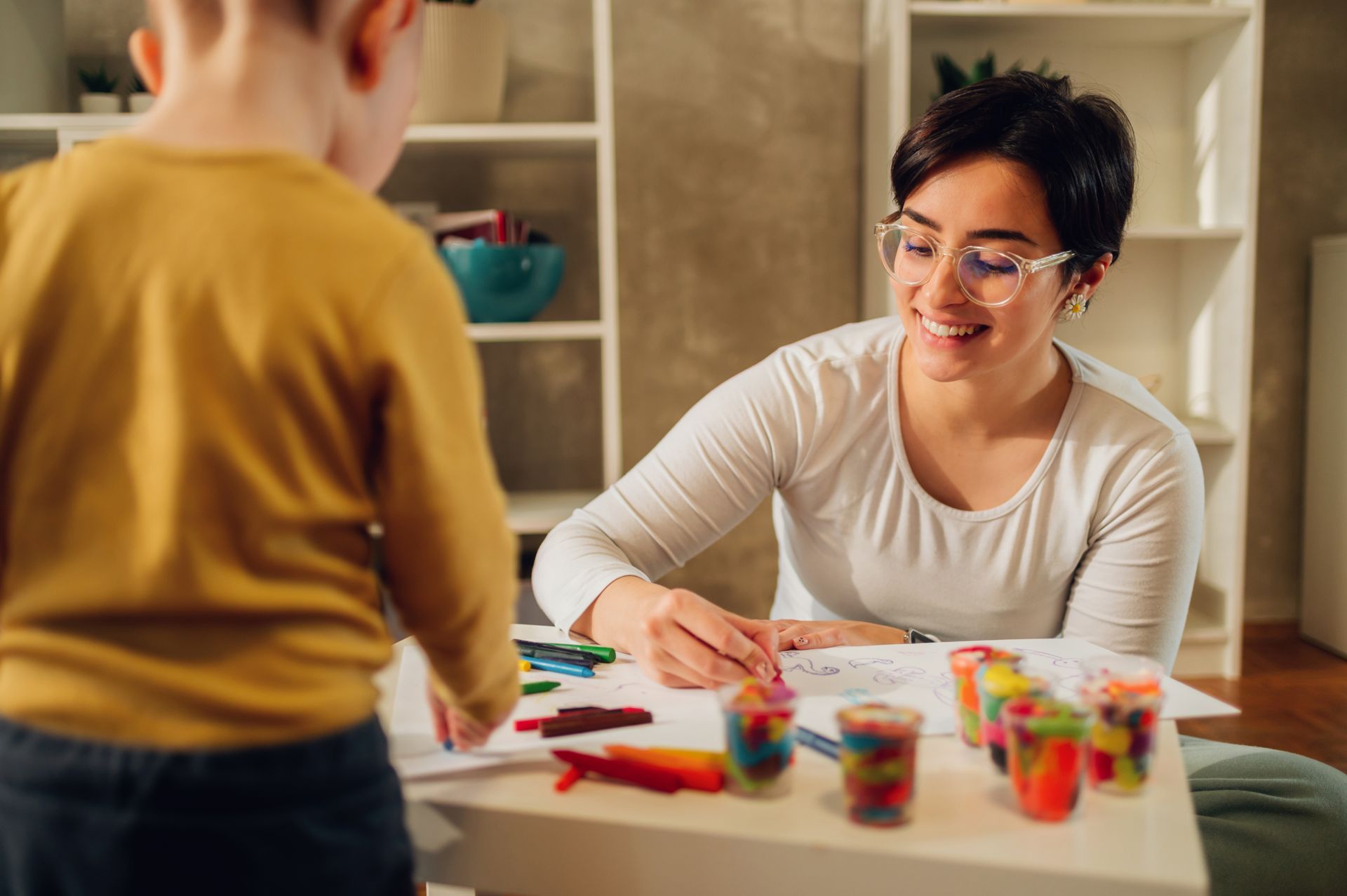 Woman and child at table with crayons and playdough; woman smiles while drawing.
