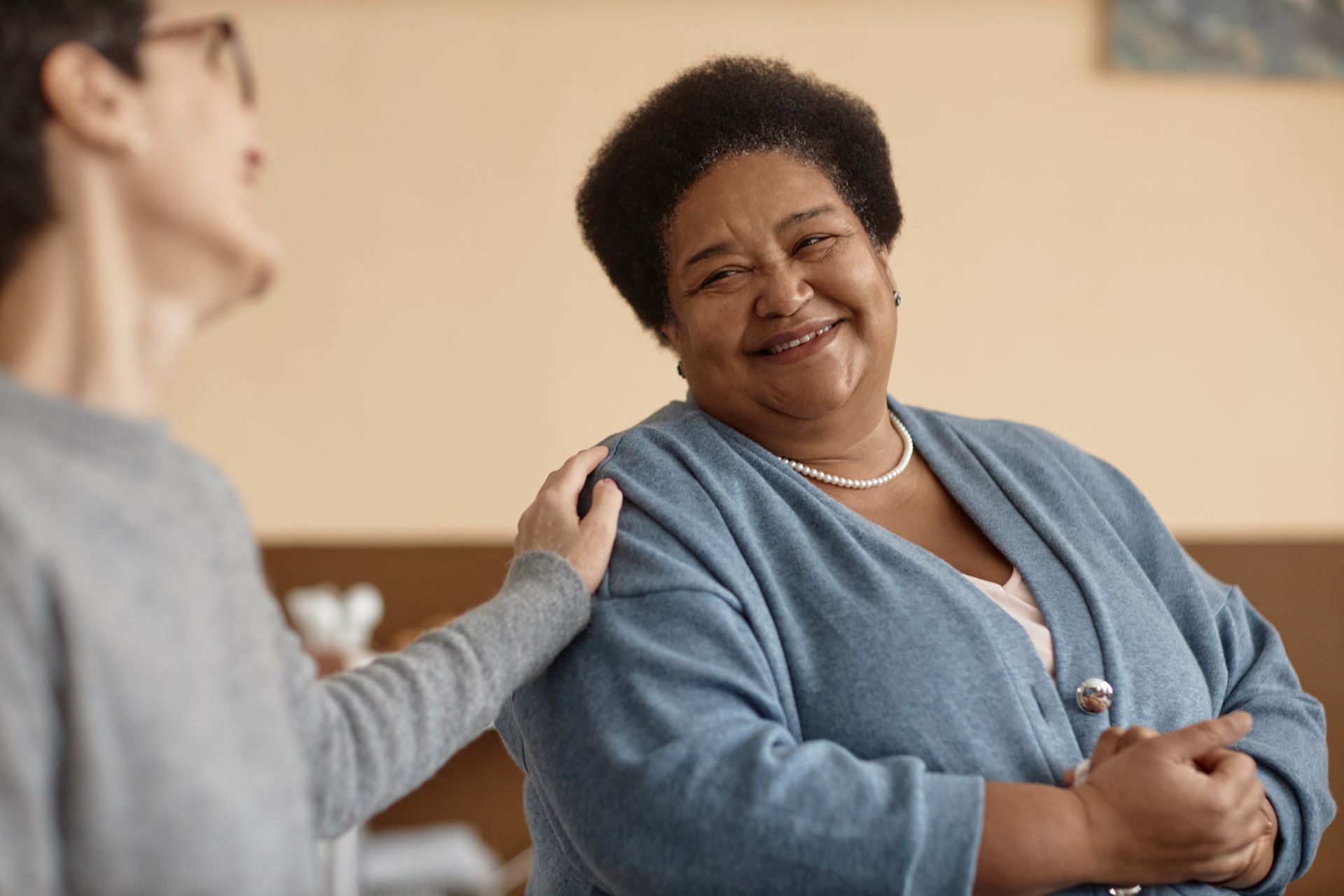 Woman in blue smiles as another person's hand rests on her shoulder. They are indoors.