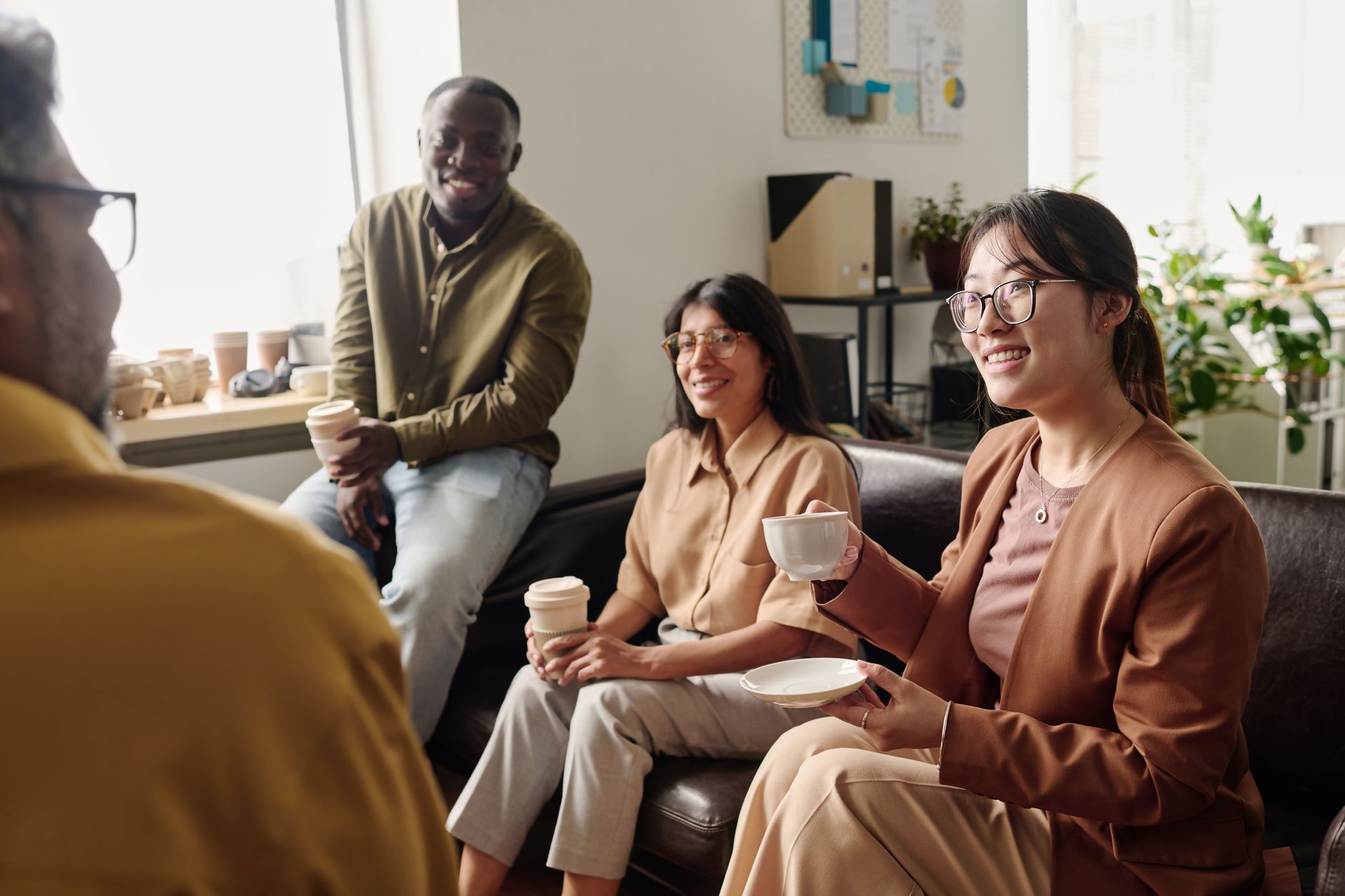 Group of people in a room, conversing while seated on a couch, holding coffee cups.