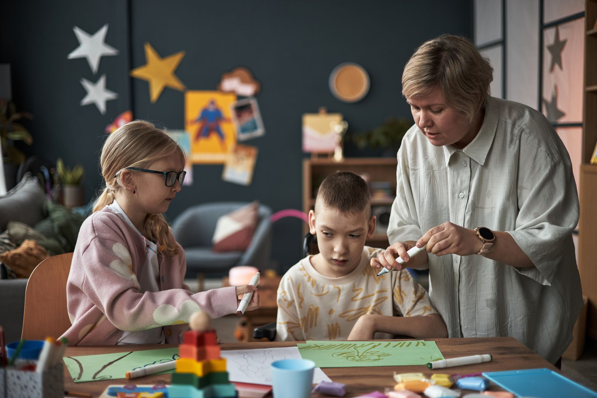 Teacher helping two children with crafts at a classroom table, with colorful supplies scattered around.