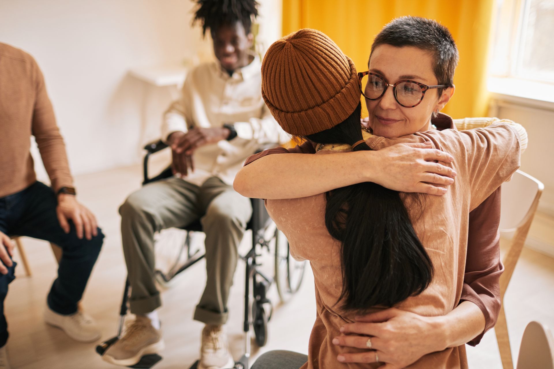 Woman in glasses hugs another woman in a beanie; other people sit in a room, one in a wheelchair.