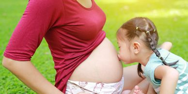 Young girl kissing the bare baby bump of a pregnant woman while sitting on grass outdoors.