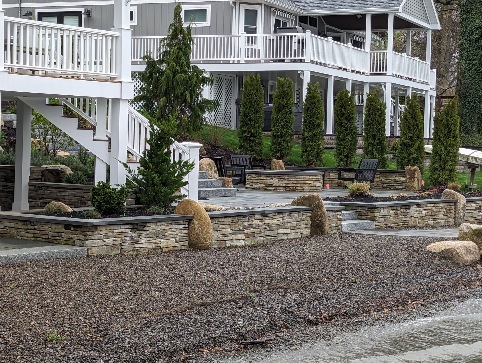 A large house with a white deck and stairs is surrounded by rocks and trees