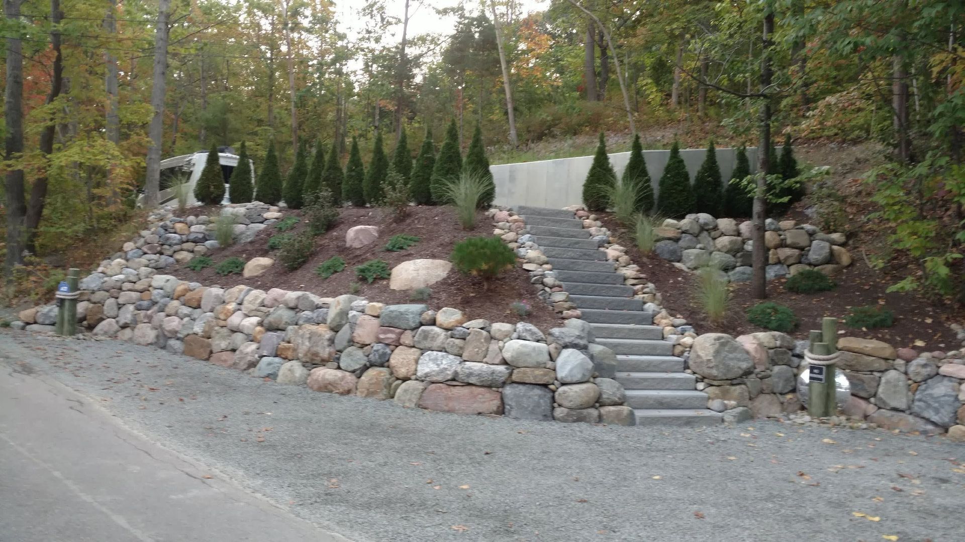 A stone wall with stairs leading up to a driveway surrounded by trees