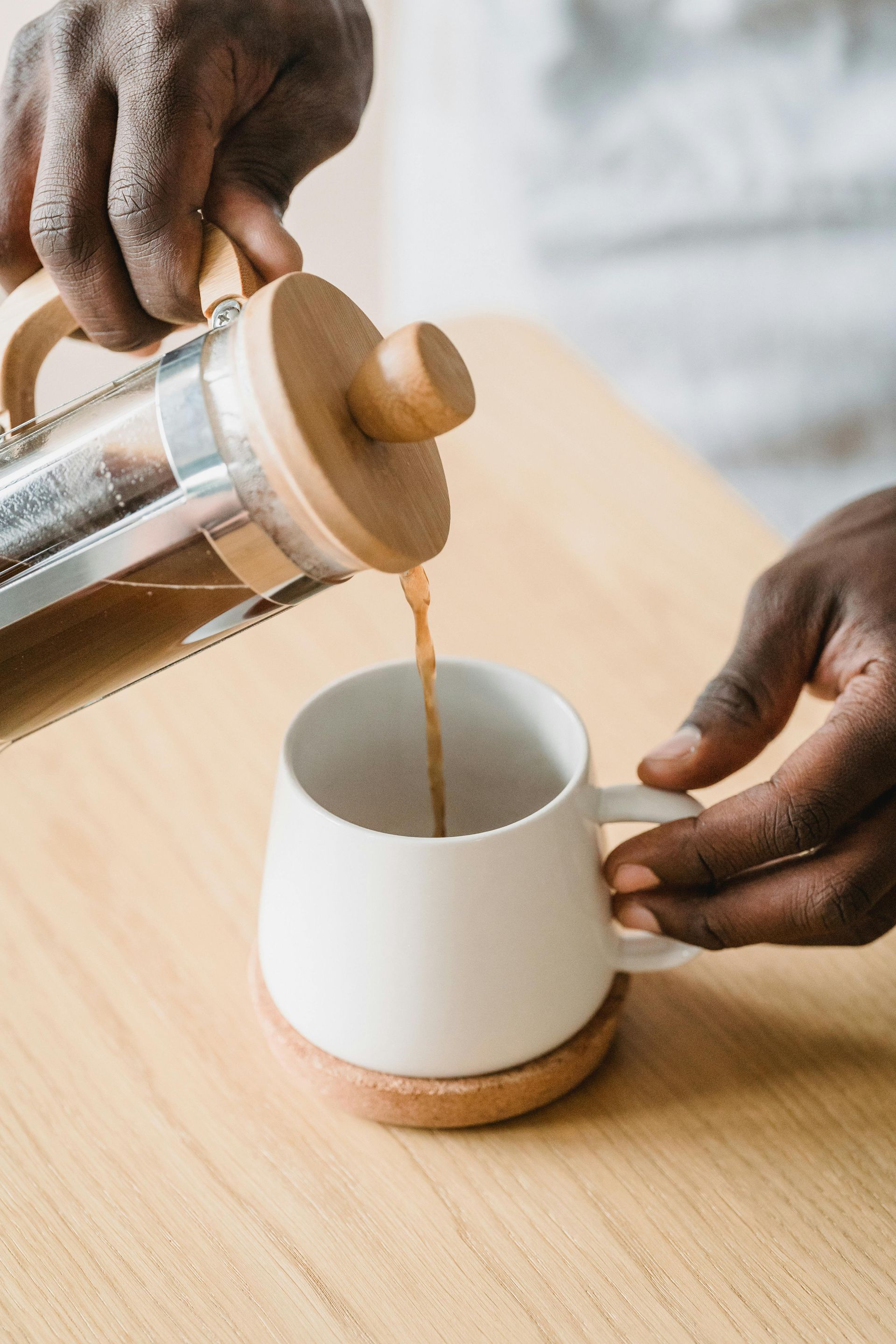 Coffee being poured from a silver and wood press into a white mug on a wooden coaster.