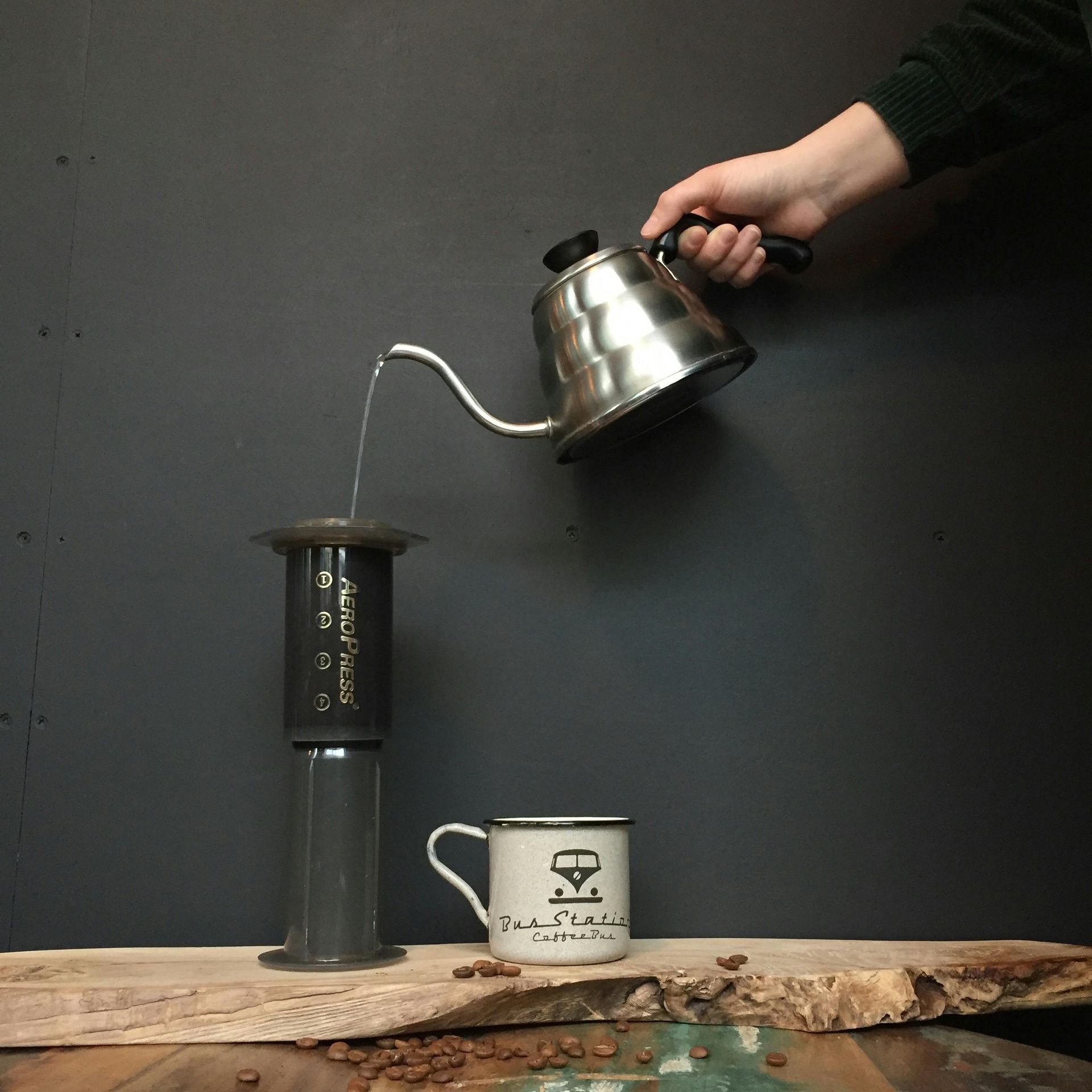 A hand pours water from a silver kettle into an AeroPress coffee maker, above a mug, on a wooden surface.