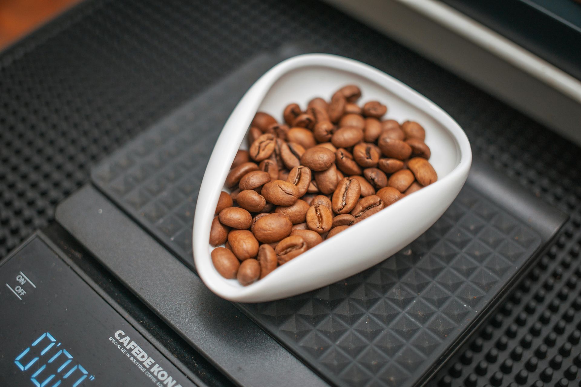 Coffee beans in a white dish on a digital scale, preparing to be weighed.