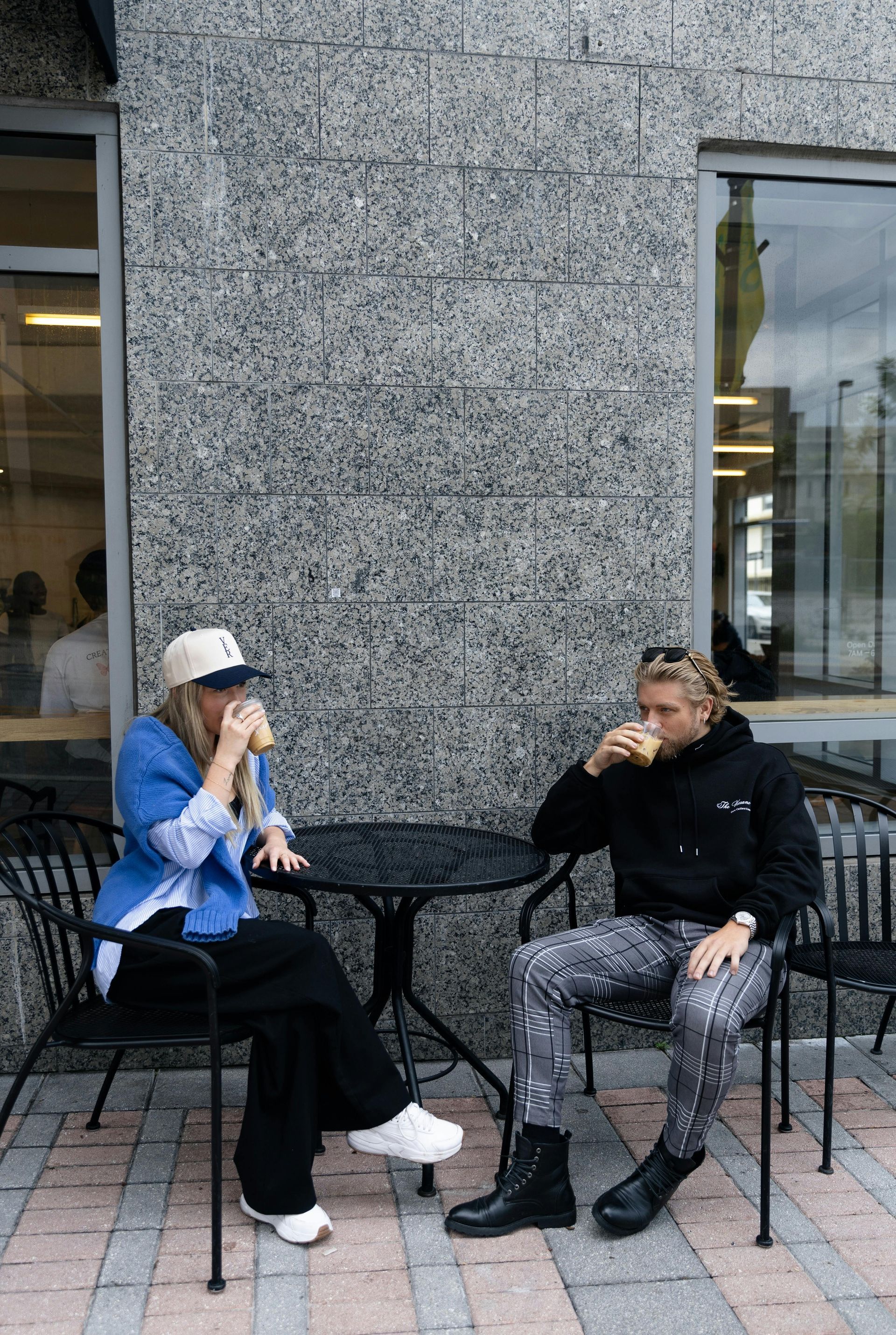 Two people sitting at a cafe table, drinking coffee. Gray stone wall in background.