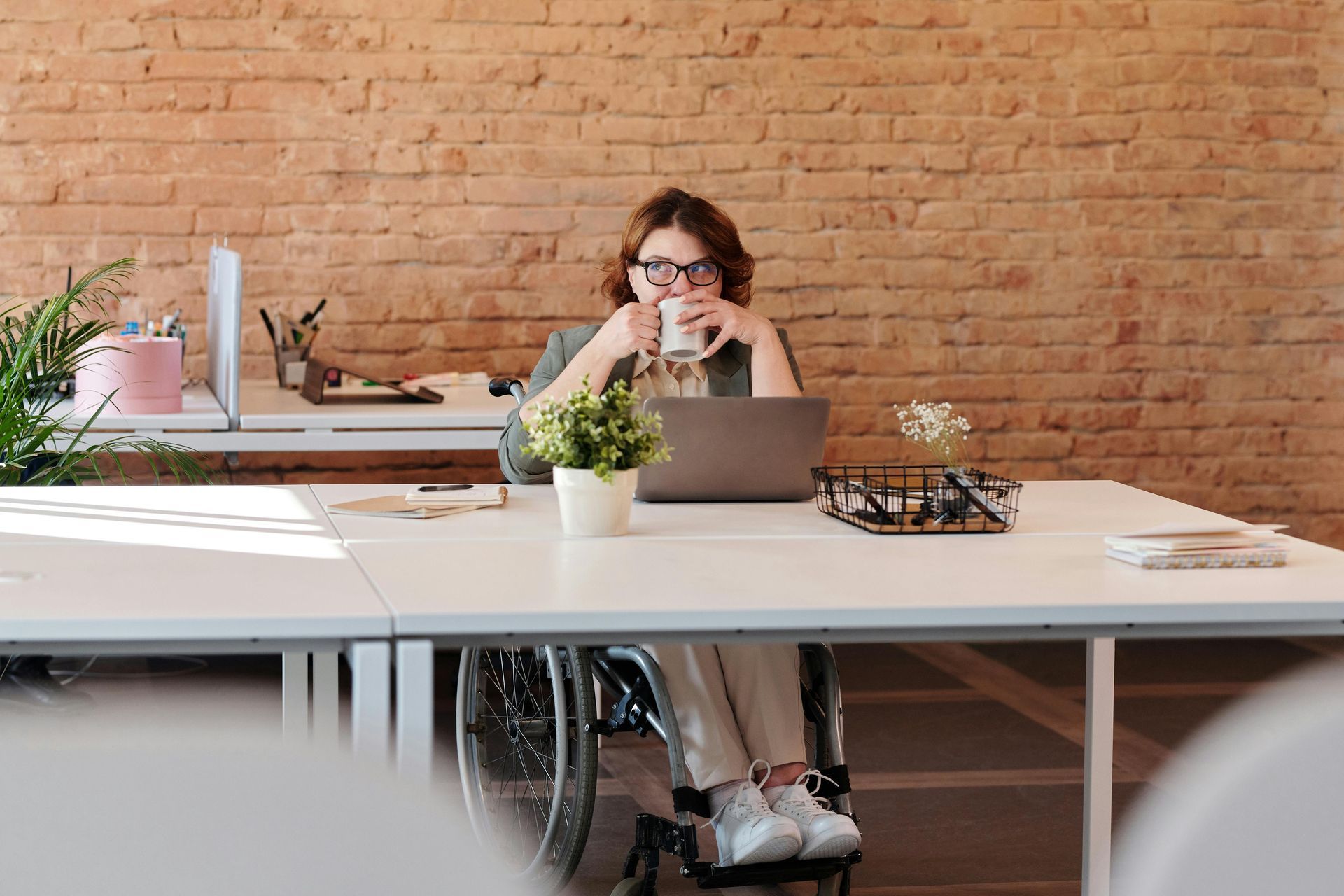 Woman in wheelchair at a desk, drinking from a mug, working on a laptop in an office setting.