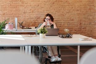 Woman in wheelchair at a desk, drinking from a mug, working on a laptop in an office setting.
