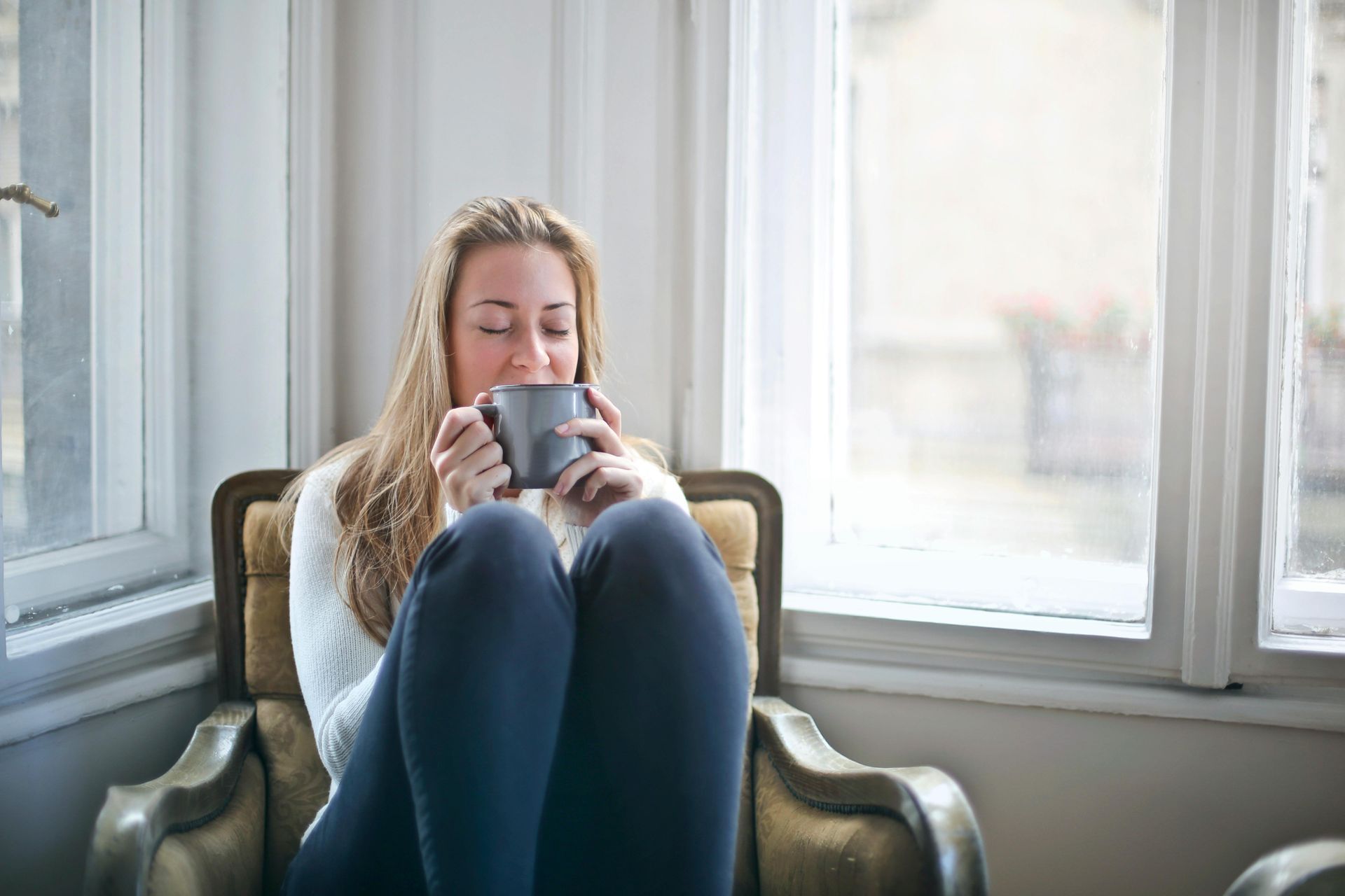 Woman seated in armchair, drinking from a gray mug near a window.