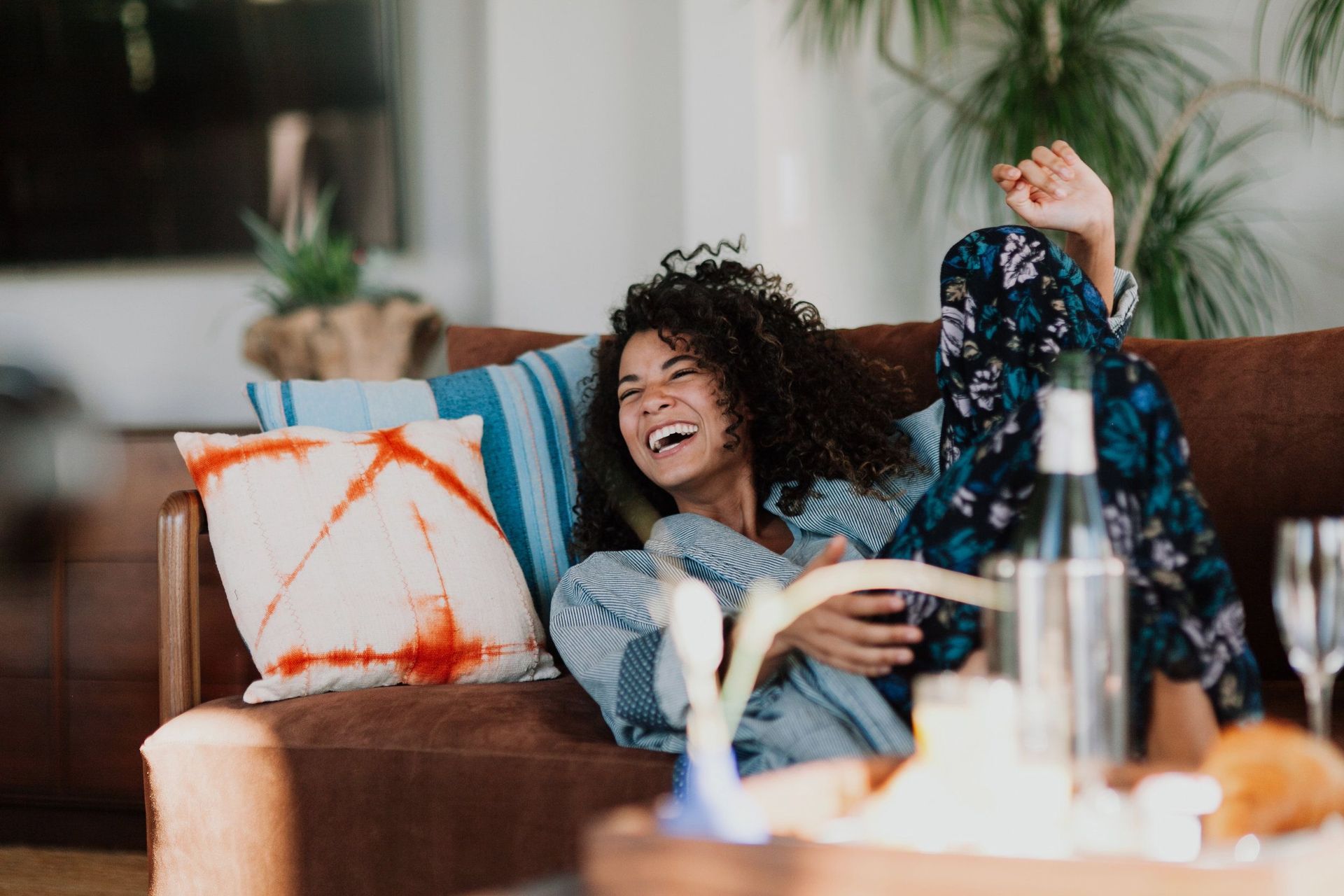 Woman laughing while reclining on a sofa, inside, next to a table with wine and snacks.