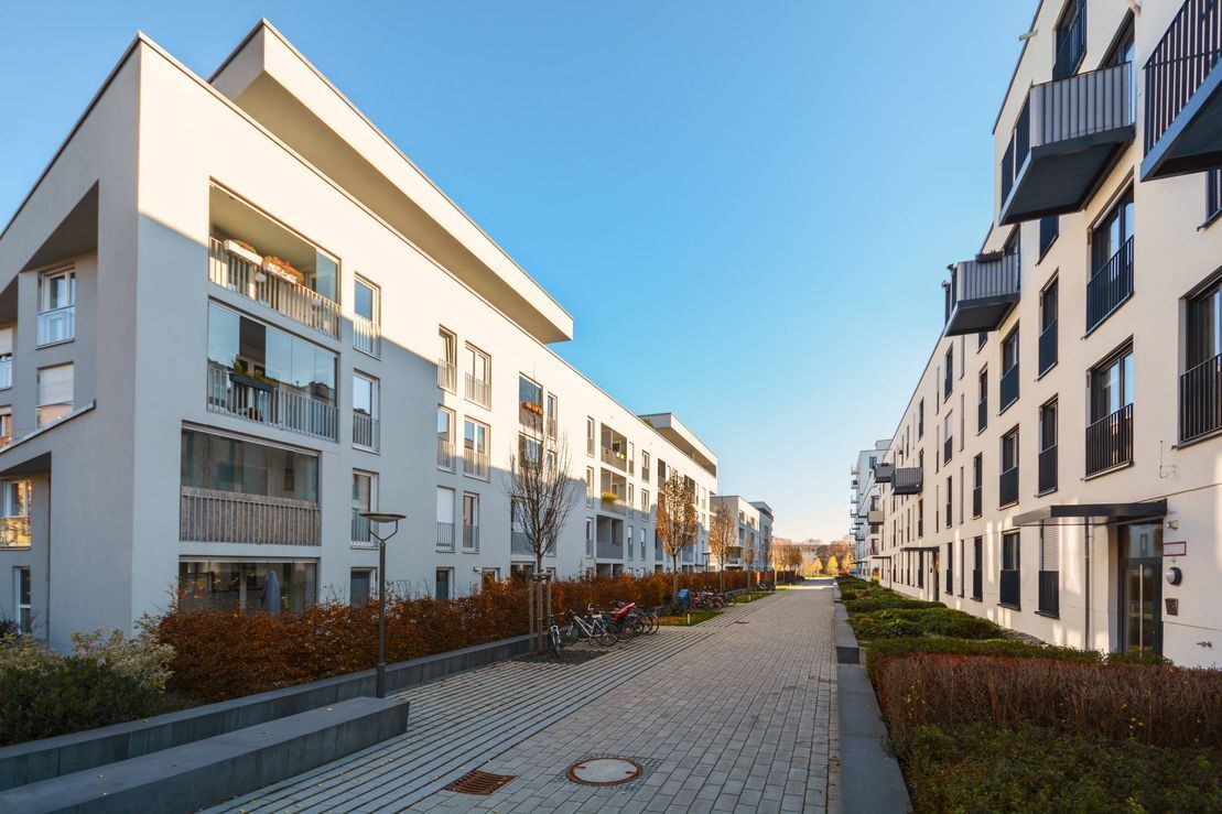 A paved walkway between two modern, multi-story white apartment buildings with balconies and landscaping.