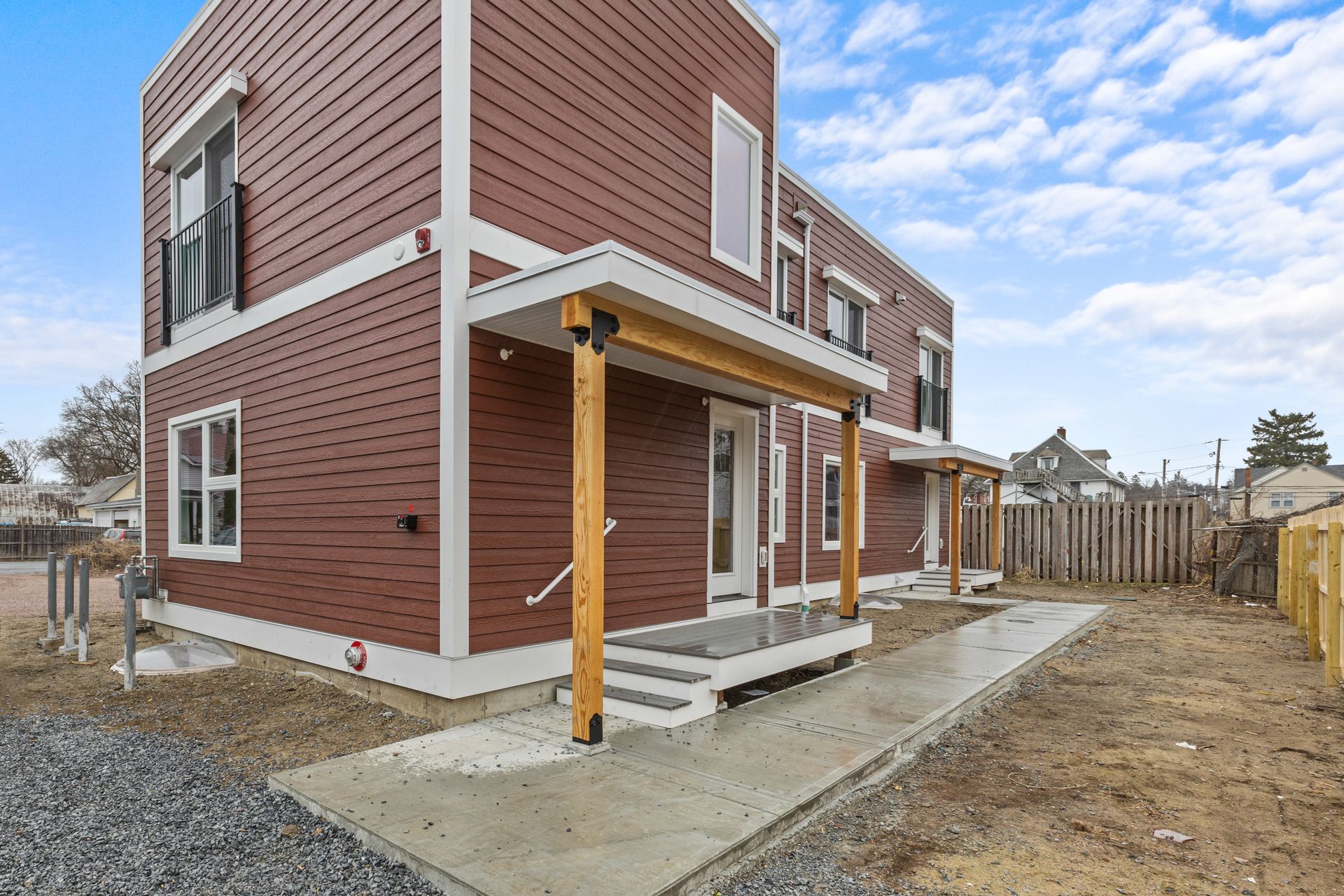 Modern two-story brown building with white trim. Porch with wooden posts. Cloudy sky, gravel, and dirt.